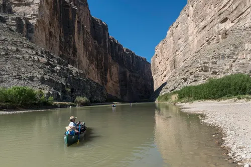 An outfitter in the stern of a canoe paddling up the Rio Grande inside Santa Elena Canyon, with more canoes strung out ahead between the canyon walls
