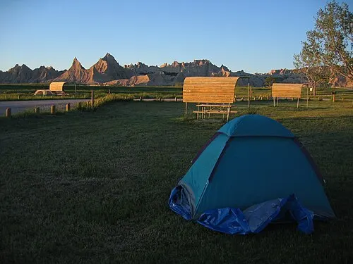 A tent pitched on the lawn at Cedar Pass Campground, with the wooden shade shelters and picnic tables and the Badlands Wall directly behind