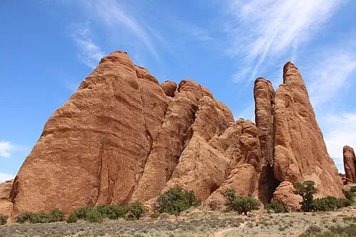 A sandy trail crosses low green shrubs toward a wall of red sandstone fins under a blue sky.