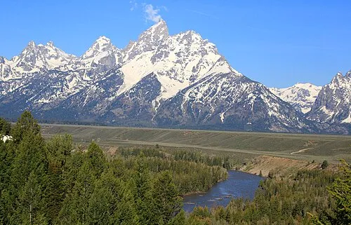 The Teton Range rising as one wall of snow-streaked peaks above a flat valley of sage and dark timber, with a bend of the Snake River in the foreground.