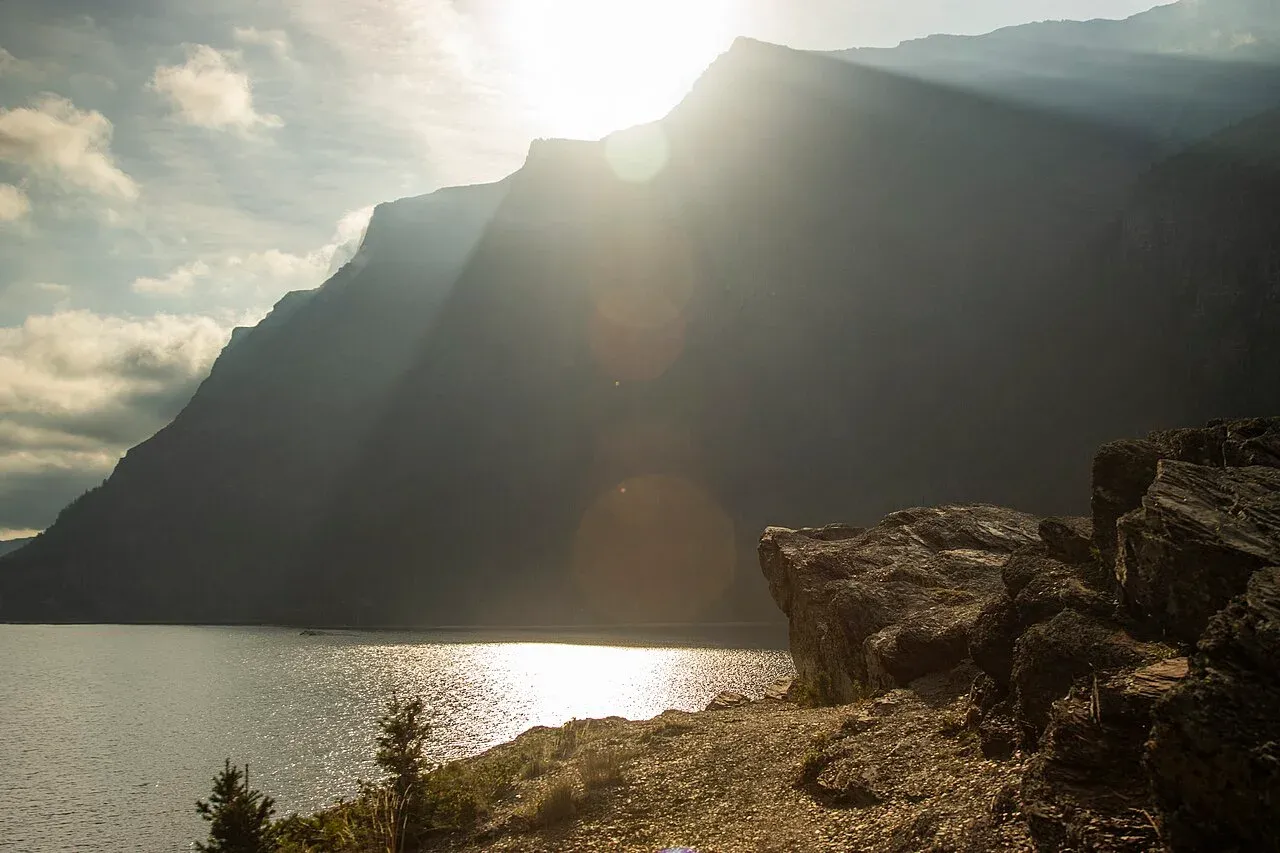 Late sun flaring over a ridge above St. Mary Lake, the rocky cliffs of Sun Point in the foreground and the lit water below.