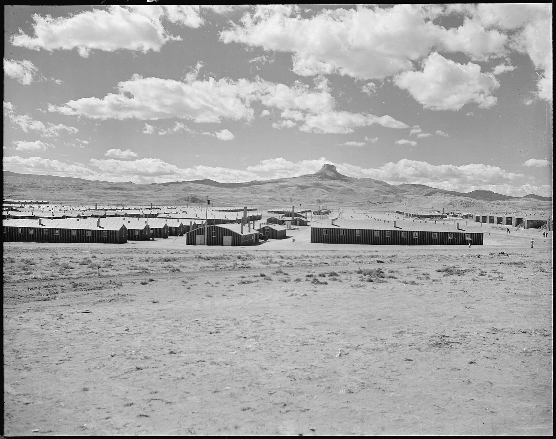Rows of tar paper barracks seen across sagebrush flats with the flat-topped summit of Heart Mountain on the horizon behind them.