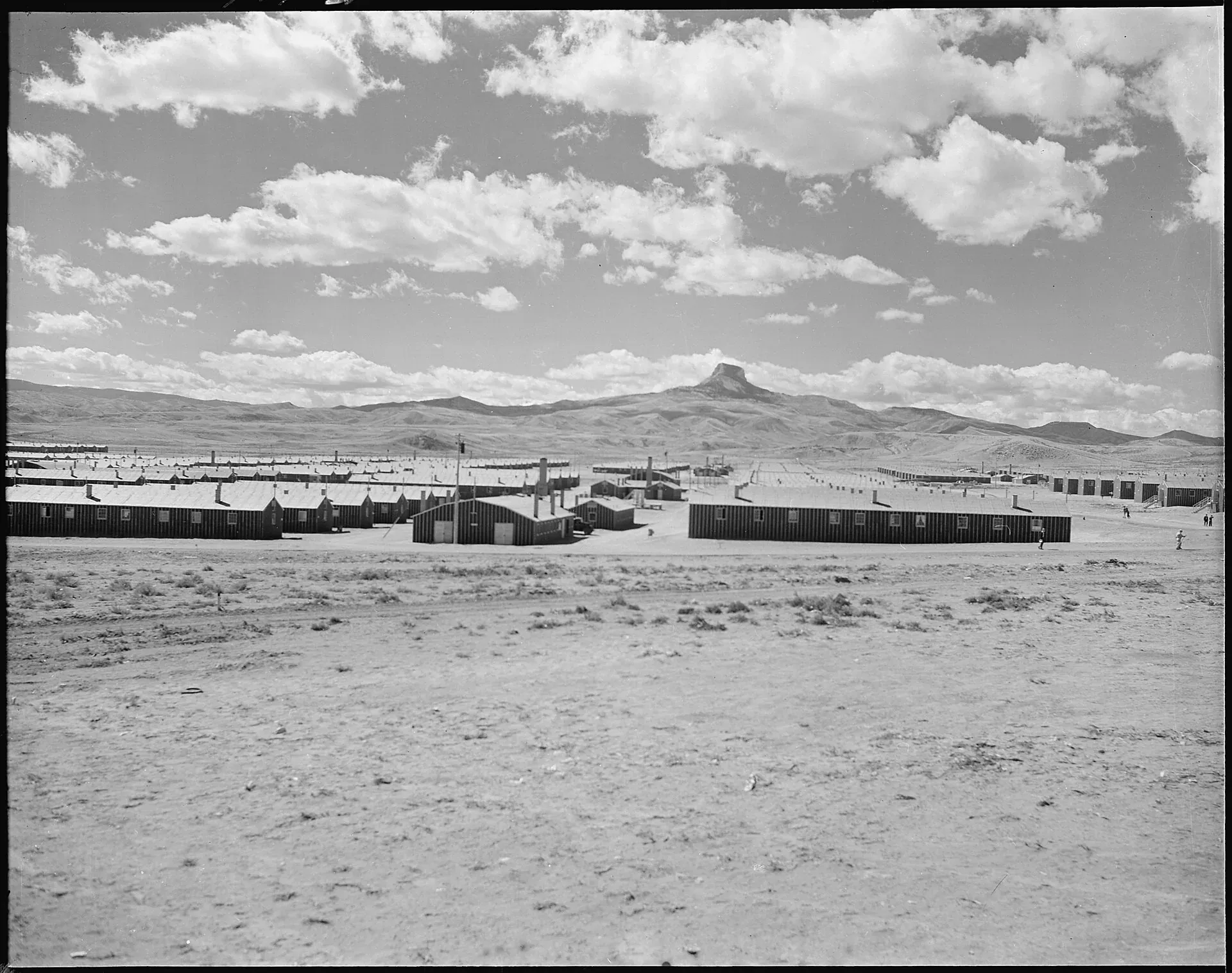 Rows of tar paper barracks seen across sagebrush flats with the flat-topped summit of Heart Mountain on the horizon behind them.