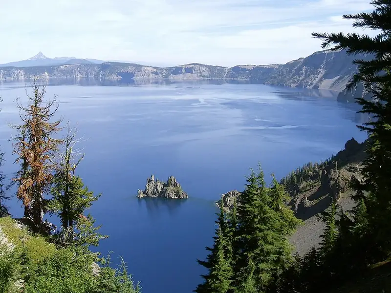 Deep blue lake water below steep caldera walls, with the rocky Phantom Ship island poking up near the shore.