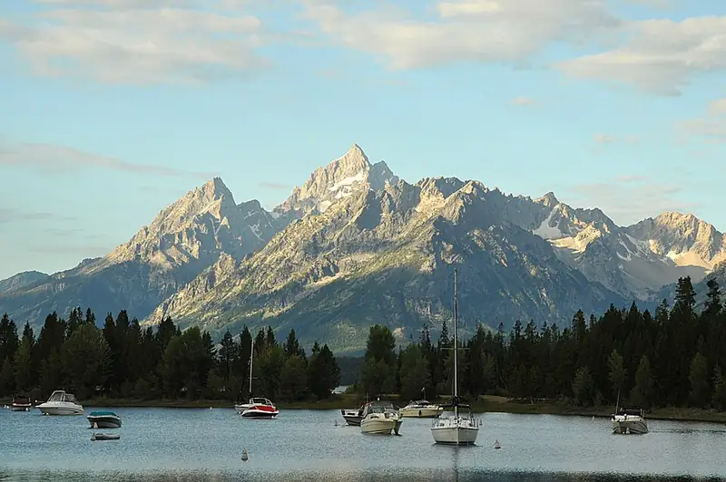 Small boats moored in a calm bay with a dark forested shoreline and the jagged Teton Range catching low evening light behind.