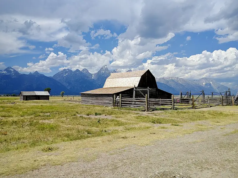 A weathered log barn with a pale shingled roof and a buck-rail corral on open sage flats, a smaller cabin off to the left and the Teton Range low on the horizon.
