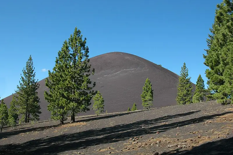 A steep, cone-shaped hill of dark cinders rises above a pine forest.