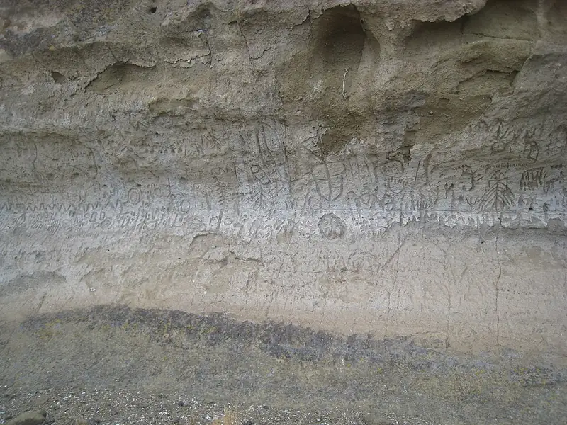 A tall pale cliff face rising above dry flat ground, where ancient carvings were cut into the rock.