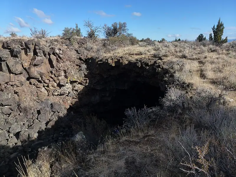 A wide, low cave entrance in dark lava rock with a metal stairway leading down into the opening.