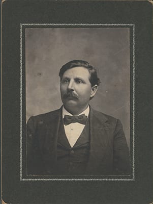 Douglas H. Johnston, the Chickasaw Nation's first appointed representative, serving from 1906 until 1939. A seated man with a full moustache and centre-parted dark hair, in a dark suit jacket and waistcoat with a white shirt and dark bow tie, turned slightly to his left and looking past the camera. A cabinet card, mounted on a dark card mount with a ruled border.